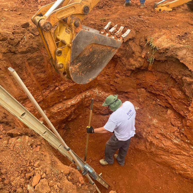 Worker digging in red clay soil with excavator and shovel nearby