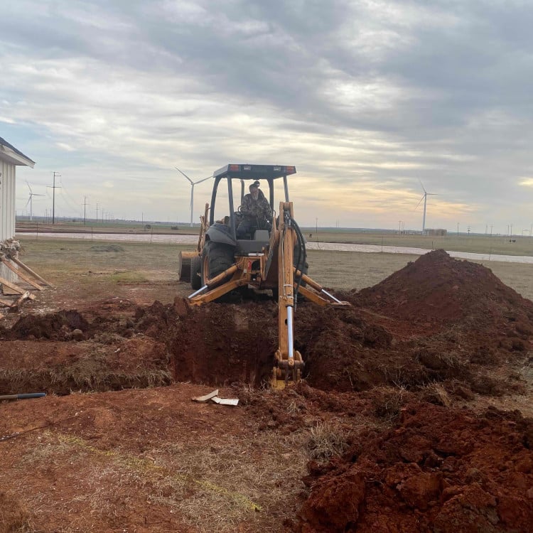 Backhoe digging hole in rural landscape with wind turbines in background